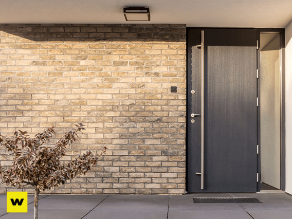 Moderne Haustür aus dunklem Metall neben heller Klinkerfassade, mit kleiner Pflanze davor.