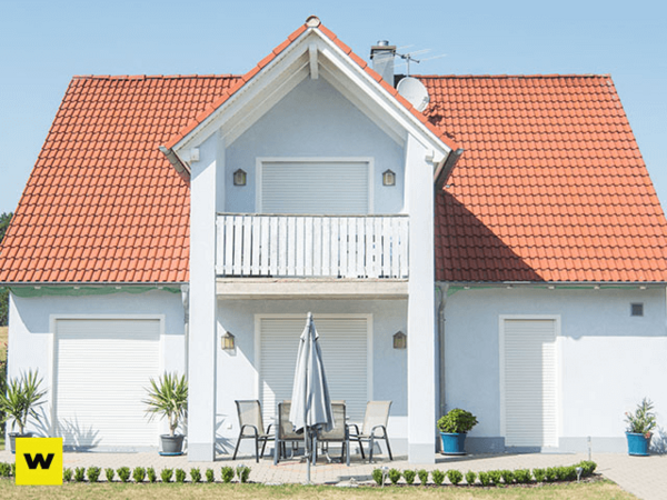 Modernes Einfamilienhaus mit rotem Dach, Balkon und Terrasse bei Sonnenschein.
